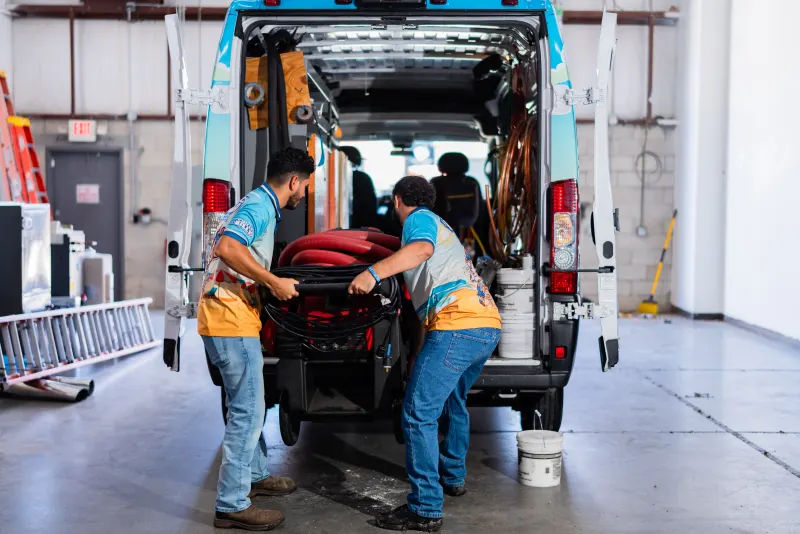 Two Chilly Billy technicians in branded shirts loading a duct-cleaning hose into the back of a service van inside the shop