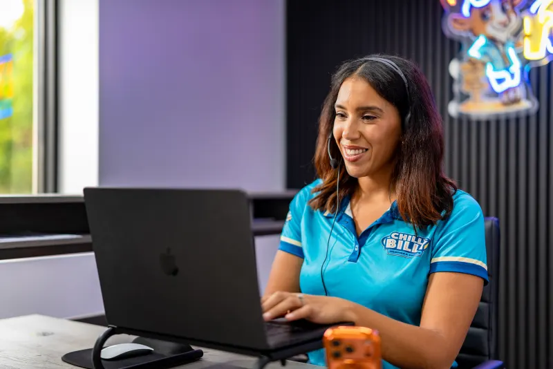 A Chilly Billy office team member in a branded polo and headset taking a call at her laptop, the Billy mascot neon sign behind her