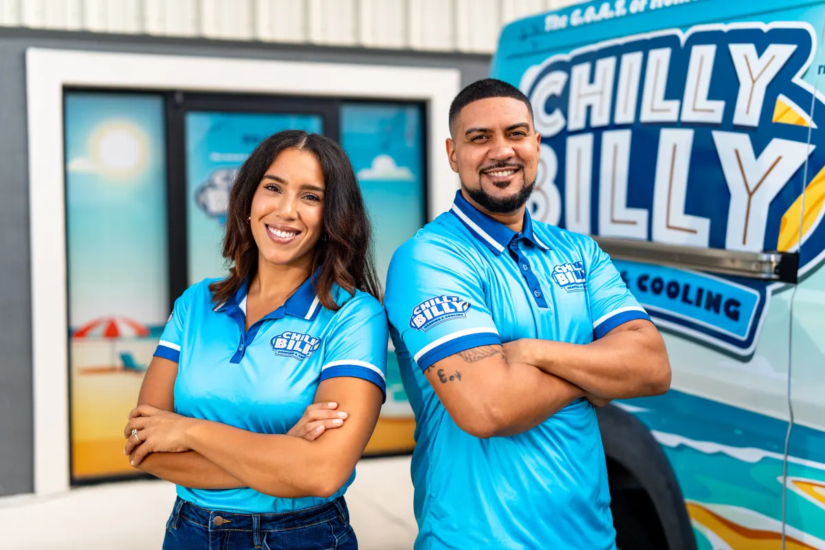 Chris and Taisha Javier, owners of Chilly Billy Heating & Cooling, arms crossed in matching blue polos beside their wrapped service van outside the Kissimmee shop