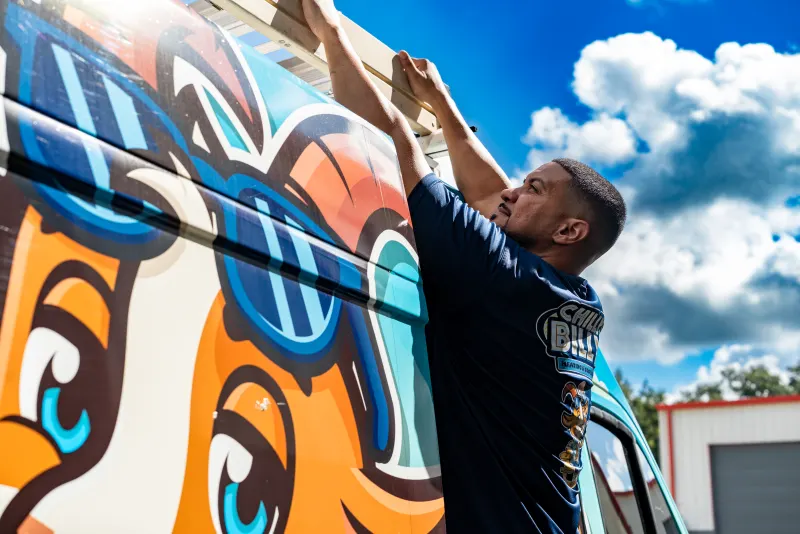 A Chilly Billy technician reaching up to secure a ladder on the roof rack of the mascot-wrapped service van under a blue sky
