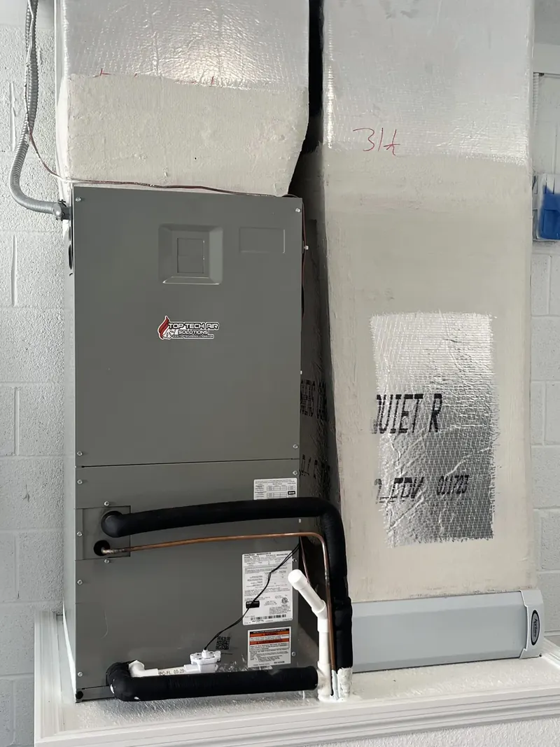 Air handler in a white utility closet with an insulated duct-board plenum above and refrigerant lines below