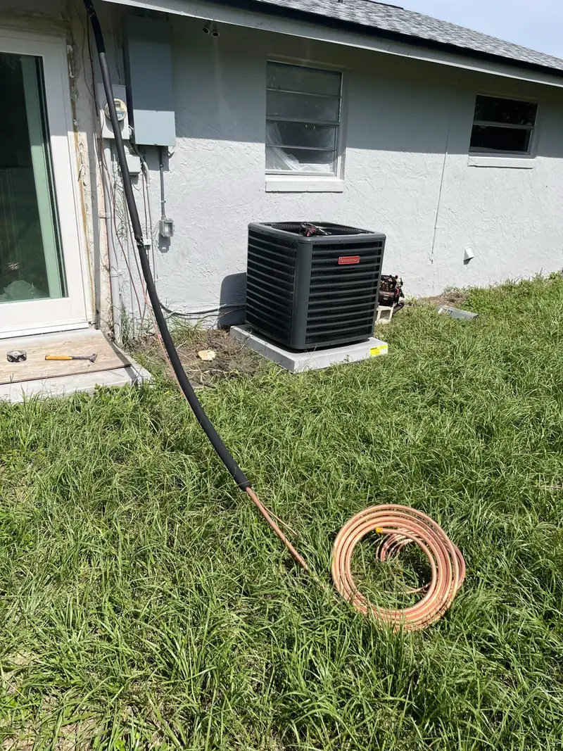 Goodman AC condenser on a pad beside a gray block home, with a coil of copper refrigerant line on the lawn